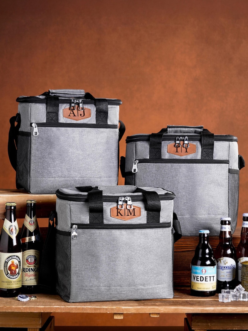 May include: Three grey insulated cooler bags with black trim and handles. Each bag features a brown leather-like patch with personalized initials: AJ, TI, and KIM. The bags have zippered compartments, side mesh pockets, and are displayed with beer bottles.