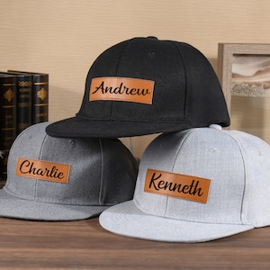 May include: Three baseball caps with personalised leather patches. The caps are black, grey, and grey. The patches have the names "Andrew", "Charlie", and "Kenneth" written in black.
