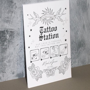 Puede incluir: Un cartel en blanco y negro con el texto "Temporary Tattoo Station" e instrucciones sobre c&oacute;mo aplicar un tatuaje temporal. El cartel tambi&eacute;n incluye el texto "Our love is forever, but this tattoo isn't." y el texto "Mark & Grace Est. 2025".