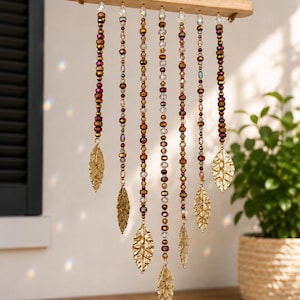 May include: A hanging mobile featuring a horizontal wooden bar suspended by a rope. Seven vertical strands of multi-colored glass beads in shades of brown, gold, and red hang from the bar, each ending with a textured gold-toned metal leaf charm.