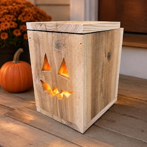 May include: A square wooden lantern featuring a carved jack-o'-lantern face with two triangular eyes and a jagged mouth. The light-colored wood box has a flat top lid and emits a warm, glowing orange light from within the hollow interior.
