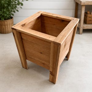 May include: A square wooden planter box with a natural light brown finish. The planter features four sturdy corner legs, a thick top rim, and flat vertical side panels. The construction shows visible wood grain and joinery details throughout.