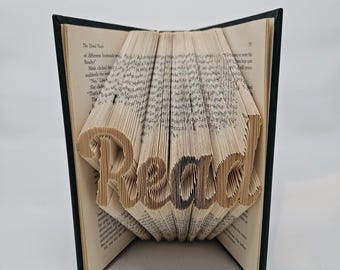 May include: A book sculpture with the word "Read" folded into the pages. The open book has a black cover and the pages are tan with printed text. The word "Read" is in a serif font.