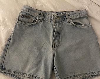 May include: Light blue denim shorts with a faded wash and a button fly. The shorts have a small pocket on the back right side and a Calvin Klein logo on the back left side.