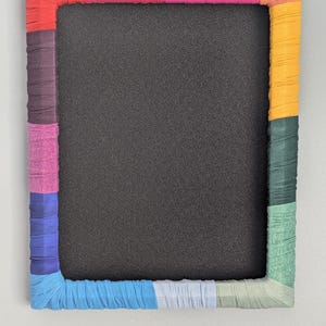 May include: A rectangular notice board featuring a black cork centre surrounded by a frame wrapped in strips of fabric in various colours including red, pink, orange, yellow, dark green, light green, light blue, royal blue, purple, and maroon.
