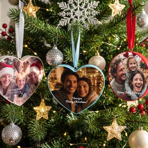 Op de afbeelding: Drie gepersonaliseerde foto-ornamenten hangen in een kerstboom. Twee zijn hartvormig met witte en lichtblauwe randen, en één is rond met een rode rand. Elk ornament toont een familiefoto en gedrukte tekst, opgehangen aan linten.