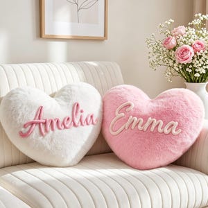 Peut inclure: Deux coussins en forme de cœur avec une texture en fausse fourrure. Un coussin est blanc avec le prénom Amelia brodé en écriture rose. L'autre coussin est rose clair avec le prénom Emma brodé en écriture rose plus clair au centre.
