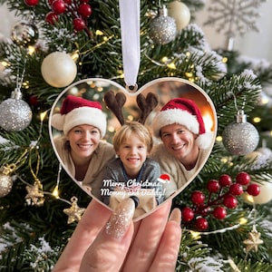 Op de afbeelding: Een hartvormige kerstversiering met een afgedrukte foto van twee volwassenen met kerstmutsen en een kind met rendiergeweien. De tekst Merry Christmas, Mommy and Daddy! staat in witte letters onderaan de versiering gedrukt.