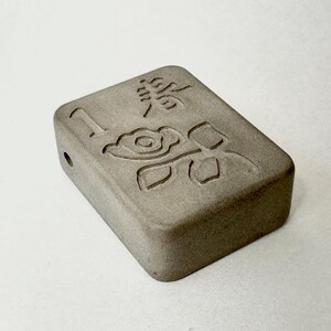 May include: A small, rectangular, gray concrete block with a carved design of a flower and leaves. The block has a small hole on one side.