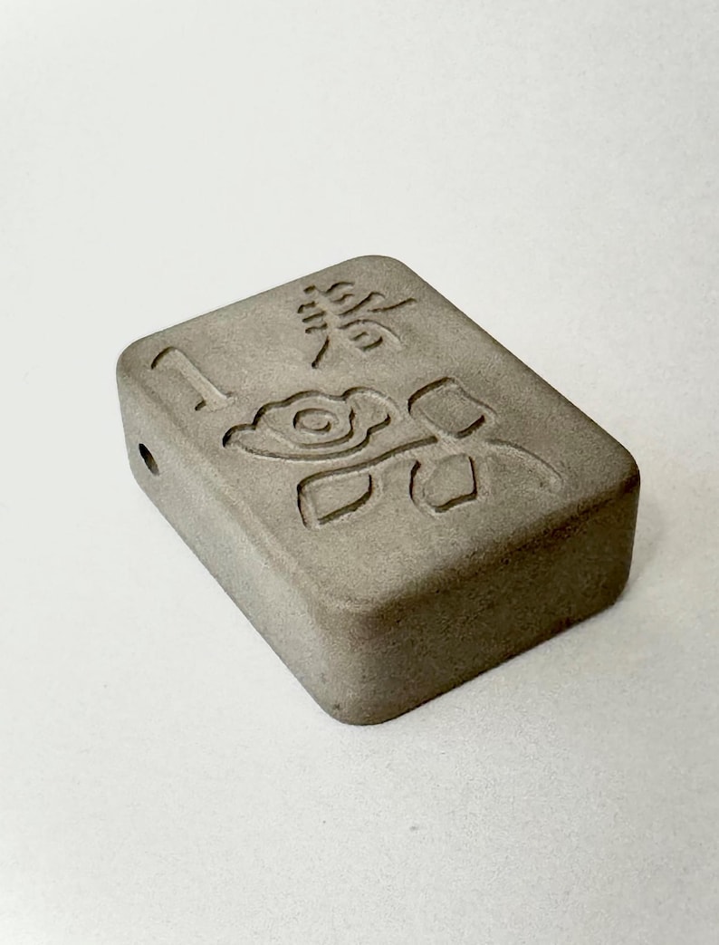 May include: A small, rectangular, gray concrete block with a carved design of a flower and leaves. The block has a small hole on one side.