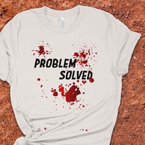 Könnte beinhalten: Weißes T-Shirt mit dem schwarzen Text "PROBLEM SOLVED", begleitet von roten Spritzgrafiken. Das T-Shirt ist am unteren Ende geknotet.