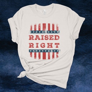 May include: Cream-colored t-shirt with a distressed American flag design. The design features red and white stripes, blue stars, and the words "RAISED RIGHT" in red block letters. The shirt is knotted at the bottom.