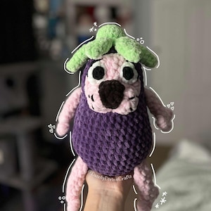Puede incluir: Un juguete de peluche de berenjena tejido a crochet con una flor verde tejida a crochet en la parte superior. El juguete tiene brazos y piernas rosas y un cuerpo morado. El juguete está siendo sostenido en una mano.