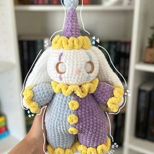 May include: A handmade crochet bunny doll in a clown costume. The plushie features a white head and ears, a purple and blue body, and yellow accents. The doll is approximately 12 inches tall.
