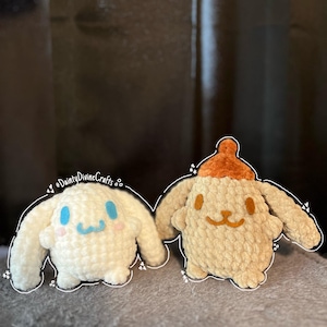 May include: Two crocheted plush toys, one white with blue eyes and a pink nose, and one tan with brown eyes and a brown hat. Both toys are shaped like a dog.