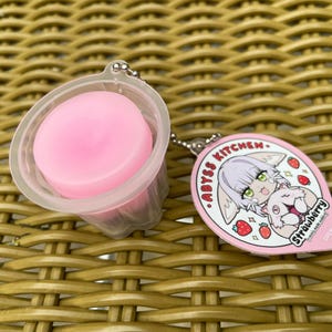 Puede incluir: Un llavero rosa y blanco con un personaje de dibujos animados con cabello rosa y un gato blanco. El llavero dice "ABYSS KITCHEN. STRAWBERRY".