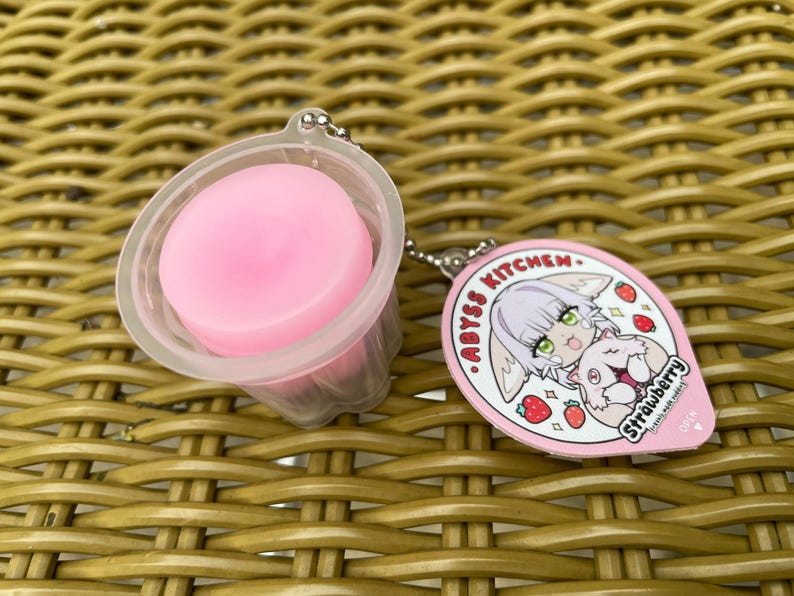 Puede incluir: Un llavero rosa y blanco con un personaje de dibujos animados con cabello rosa y un gato blanco. El llavero dice "ABYSS KITCHEN. STRAWBERRY".