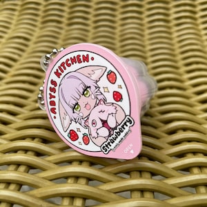Puede incluir: Llavero rosa y blanco con un personaje de dibujos animados sosteniendo una fresa. El llavero dice "Abyss Kitchen. Strawberry" y tiene una peque&ntilde;a flecha abierta.