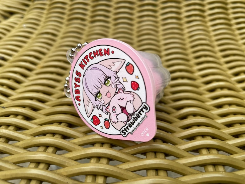 Puede incluir: Llavero rosa y blanco con un personaje de dibujos animados sosteniendo una fresa. El llavero dice "Abyss Kitchen. Strawberry" y tiene una peque&ntilde;a flecha abierta.