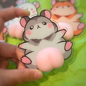 May include: A selection of cartoon hamster-shaped acrylic charms. The charms feature a grey, white, and pink hamster design with a pink peach-shaped bottom. The background is a bright green colour.
