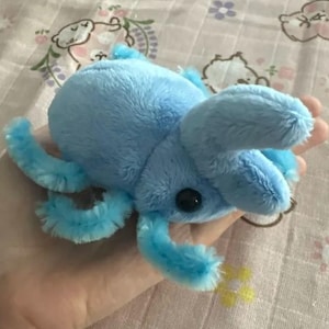 May include: A light blue plush beetle toy with a soft, fuzzy texture. The insect features a rounded body, six articulated legs, and two prominent, curved horns extending from the head. It has a single visible black, glossy, round eye.