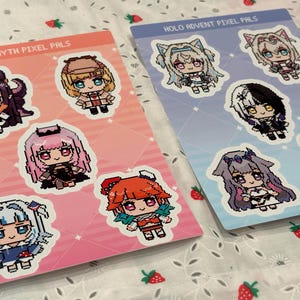 May include: Two sticker sheets featuring pixel art characters. One sheet has a pink and orange gradient background with the text "HOLOMYTH PIXEL PALS". The other sheet has a blue gradient background with the text "HOLO ADVENT PIXEL PALS". Each sheet has several character stickers.