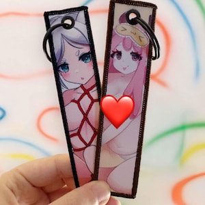 Puede incluir: Dos marcapáginas rectangulares de estilo anime. Uno presenta un personaje de pelo blanco con ojos azules y detalles de cuerda roja. El otro muestra un personaje de pelo rosa con una máscara para dormir. Ambos tienen anillos metálicos negros.