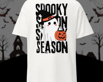 May include: White t-shirt with a Halloween-themed graphic. The design features a cartoon ghost wearing a witch hat and holding an orange pumpkin. The text "Spooky Season" is printed in black.