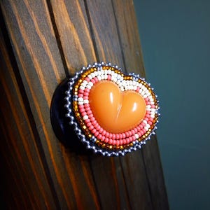 May include: A heart-shaped accessory featuring a smooth, solid peach-colored center surrounded by concentric rings of white, pink, and amber-colored seed beads, finished with an outer border of metallic silver beads on a black circular base.