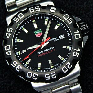 May include: A TAG Heuer Formula 1 watch with a black dial and a stainless steel band. The watch features a black bezel with white numerals and a red second hand. The watch face displays the TAG Heuer logo and the words "Formula 1".