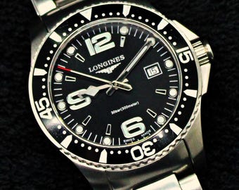 May include: A stainless steel Longines wristwatch with a black dial, a rotating bezel, and a metal link bracelet. The watch face features large white numerals at 6 and 12 o'clock, a date window at 3 o'clock, and text reading 30bar(300meter) and SWISS MADE.