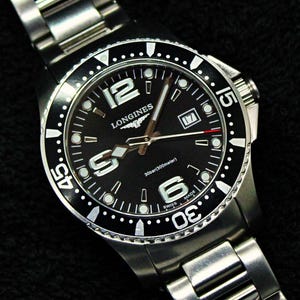 Peut inclure: Une montre-bracelet Longines en acier inoxydable avec un cadran noir, dotée de grands chiffres argentés à 6 et 12 heures, d'un guichet de date à 3 heures et d'une lunette noire rotative. Le cadran affiche Longines, 30bar(300meter) et Swiss Made.