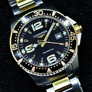 May include: A two-tone stainless steel and gold-tone wristwatch featuring a black dial with the Longines logo, large Arabic numerals at 6, 9, and 12, a date window at 3, luminous hands, and a rotating black bezel with gold-tone markers and 30bar depth rating.