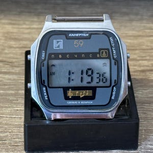 May include: A retro digital wristwatch with a silver case and a rectangular face. The watch face is dark blue with white text and shows the time as 1:19:36 AM. The watch has several buttons and the text "KAMERTON" is visible.