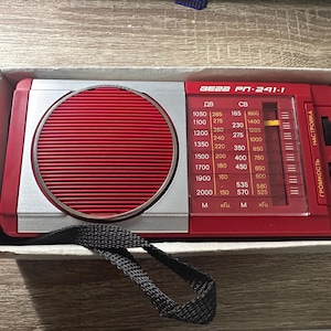 May include: A vintage red and silver radio, model BEZA PN-241-1, with a round speaker and a tuning dial. The radio is in its original cardboard box with a black strap. The radio's dial displays frequency numbers.