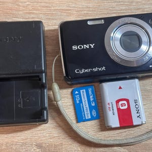 May include: A black Sony Cyber-shot digital camera with a 12.1 megapixel lens, a black battery charger with a charge indicator light, a blue SanDisk 512MB Memory Stick Pro Duo card, and a white and red Sony NP-BG1 lithium ion G-type battery.