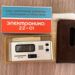 May include: An Elektronika 22-01 digital travel alarm clock with a white face, black buttons, and a digital display showing 3:05. It includes a blue and red cardboard box with Cyrillic text, a brown velvet protective pouch, and paper documentation.