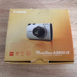 May include: A tan cardboard box for a silver Canon PowerShot A3300 IS digital camera. The box features the Canon logo, the camera model name, and technical specifications including 16.0 megapixels, 5x zoom, 28mm lens, and a 3.0 inch LCD screen.