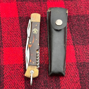 May include: A vintage pocket knife with a brown handle and multiple blades, including a corkscrew. It is next to a black leather sheath with a silver snap. The knife has a gold-colored metal end and a key ring attachment.