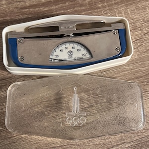 Könnte beinhalten: Vintage Metallwaage mit blau-silbernem Zifferblatt, untergebracht in einem weißen Kunststoffgehäuse. Die Waage hat eine gebogene Anzeige mit Zahlen und eine transparente Kunststoffabdeckung mit einem olympischen Logo.