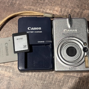 May include: A silver Canon PowerShot SD200 digital camera with a Canon battery charger, a 32 Mbyte memory card, and a Canon battery. The camera has a zoom lens and a wrist strap.
