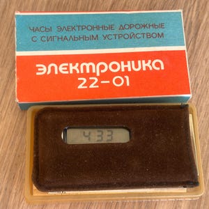 May include: A vintage Elektronika 22-01 digital travel clock with an alarm feature, housed in a brown velvet-textured case. The clock displays the time 4:33 on a small digital screen. It is presented with its original blue, white, and red box.