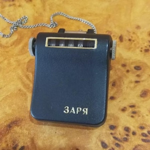 May include: A black mechanical counter with a silver chain. The counter has a rectangular window displaying numbers and the Cyrillic word "Zarya" printed on the front. The counter is on a wood-grain surface.