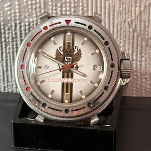 May include: A silver wristwatch with a white face, date display, and the word "COMMANDOS". The watch has a black bezel with red accents and a detailed emblem. The watch is set on a black stand.