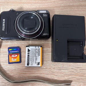 Può includere: Una fotocamera digitale Fujifilm nera con obiettivo zoom 10x e 14 megapixel, accompagnata da una scheda di memoria SD Transcend da 1 GB, una batteria agli ioni di litio Fujifilm NP-45A e un caricabatterie Olympus nero con spia gialla.