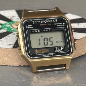 Pode incluir: Um relógio de pulso digital dourado com um mostrador retangular apresentando o texto ELEKTRONIKA 5 em cirílico. O ecrã preto mostra a hora 1:05 e inclui ícones de música, um sinal e uma figura a correr, com botões laterais.