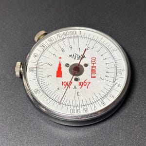 May include: A vintage, circular instrument with a white face, featuring a red star and the numbers 0-90. The dial includes the text "1917 1967" and "OKTASPB". The instrument has two silver knobs and a needle.