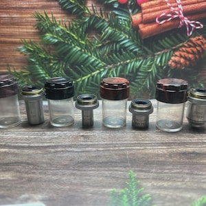 May include: A collection of small glass vials with dark brown and black caps, and silver-colored metal components. The vials are arranged in a row against a wooden background with Christmas decorations.