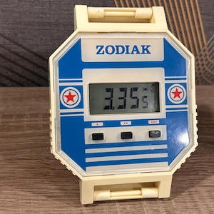 May include: Vintage ZODIAK digital watch with a cream-coloured case and a blue and white face. The watch displays the time 3:35. Features include a red star emblem and the ZODIAK logo.
