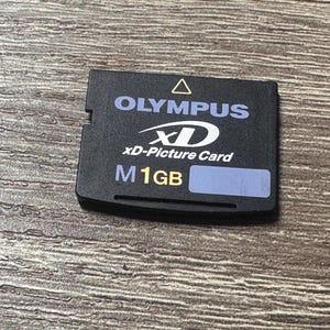 Puede incluir: Tarjeta xD-Picture Olympus negra con texto blanco y azul. La tarjeta dice "OLYMPUS", "xD-Picture Card" y "M 1GB". Una pequeña pegatina azul está en la parte inferior derecha. La tarjeta está sobre una superficie de madera.